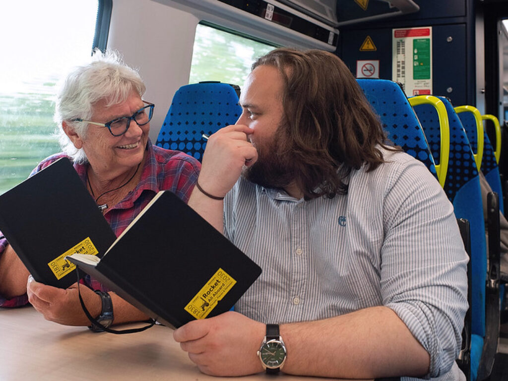 A man and woman sat on a train talking to each other holding books with the rocket all aboard logo on it.
