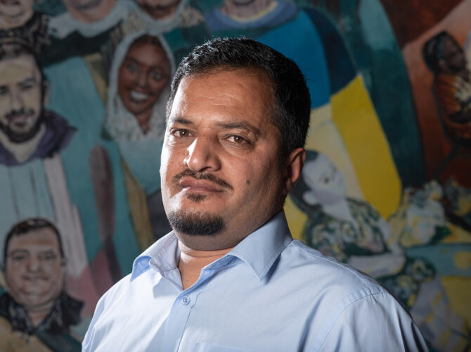 Farid Ahmed Hiwa Syd Sharif Chapashi standing in front of the colourful community artwork at Edge Hill Station.